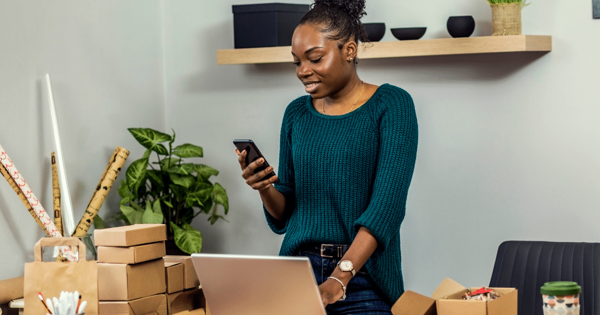 Woman in a green sweater using a smartphone in a small business setting with packages and a laptop, exploring bad credit loan options.