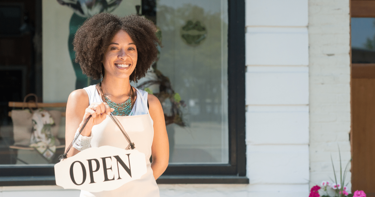 Smiling woman holding an 'OPEN' sign outside her business, representing women entrepreneurs and unsecured credit options.