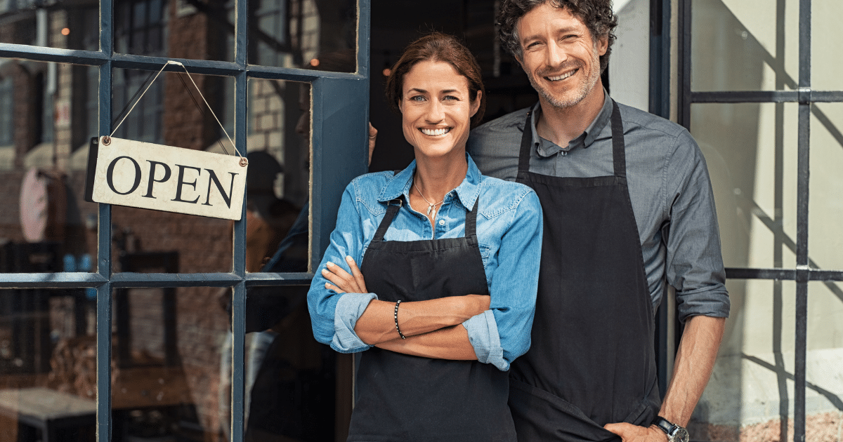 Smiling grocery business owners standing by an open sign, representing bad credit business loans for growth and cash flow.
